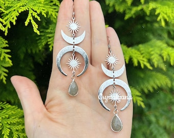 Può includere: Orecchini argentati con un design celeste, con lune crescenti, scoppi di stelle e gemme a forma di goccia. Gli orecchini sono mostrati su uno sfondo di fogliame verde, evidenziando il loro design.