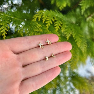 Puede incluir: Dos pares de pendientes de oro: en forma de estrella con detalles de cristal transparente y en forma de media luna. Los pendientes se presentan sobre un fondo de follaje verde.