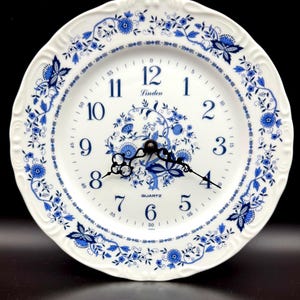 May include: A round white ceramic wall clock featuring a blue floral pattern around the scalloped edge and center. The clock face displays blue Arabic numerals from 1 to 12, the word Linden, the word QUARTZ, and black ornate metal clock hands.