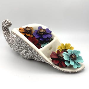 May include: A ceramic cornucopia-shaped bowl with a speckled gray and white exterior and a smooth cream interior. The bowl is filled with several small, painted pine cones in vibrant shades of orange, purple, red, yellow, and turquoise blue.