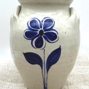 May include: A cream-colored ceramic crock with a wide rim and two side handles. The front features a hand-painted dark blue flower with a central dot and two leaves on a single stem. The surface has a speckled texture throughout the glaze.