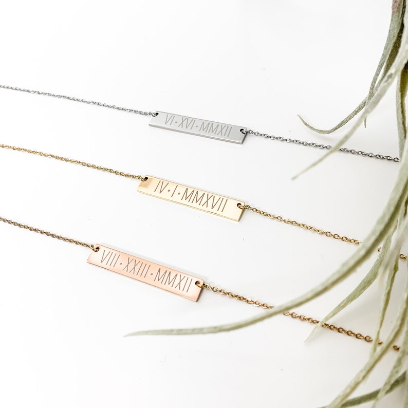 May include: Three gold bar necklaces with Roman numeral dates engraved on each. The top necklace is silver, the middle necklace is gold, and the bottom necklace is rose gold.