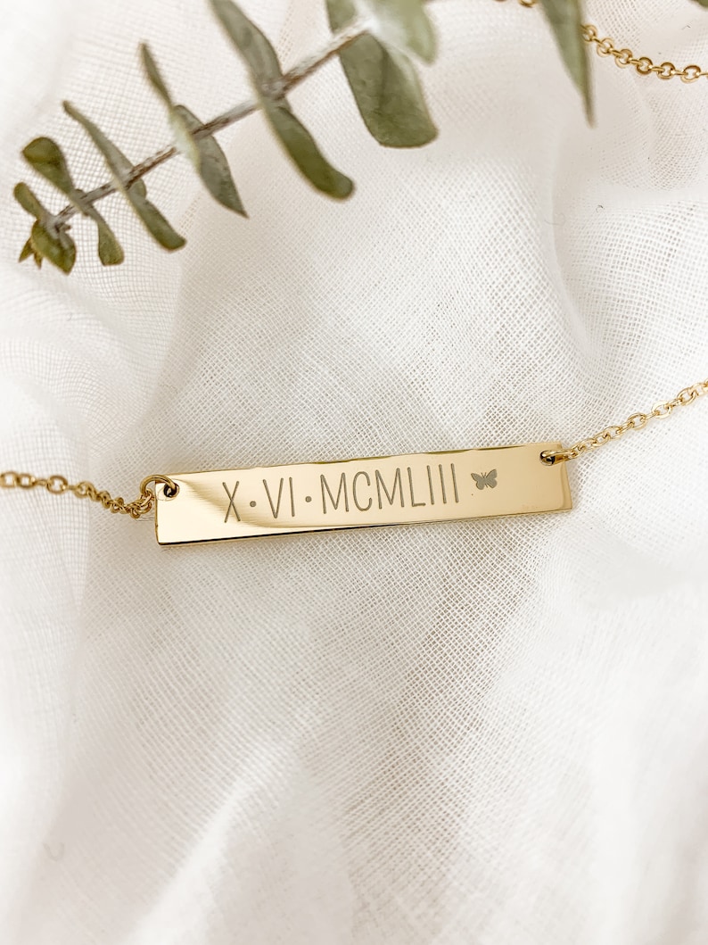 May include: Gold bar necklace with a butterfly charm and the inscription "X.VI. MCMLI" on a white fabric background.