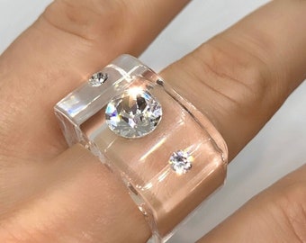 May include: A wide, clear resin ring featuring a smooth, nude-coloured interior band. The exterior is embedded with three round, faceted clear crystals of varying sizes, arranged in a diagonal pattern across the top surface of the band.