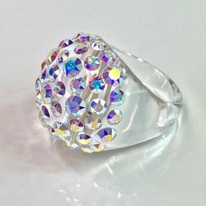 May include: A clear, transparent ring featuring a large, rounded dome top embedded with numerous iridescent, multi-coloured crystals that reflect light in shades of blue, yellow, and pink. The band is smooth, thick, and made of clear material.