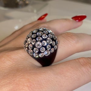May include: A black ring with a large, round, clear crystal top. The ring is set with many small, clear crystals.