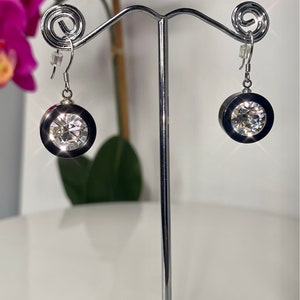 May include: A pair of silver earrings with a large, clear crystal stone set in a black bezel. The earrings are hanging from a silver stand.