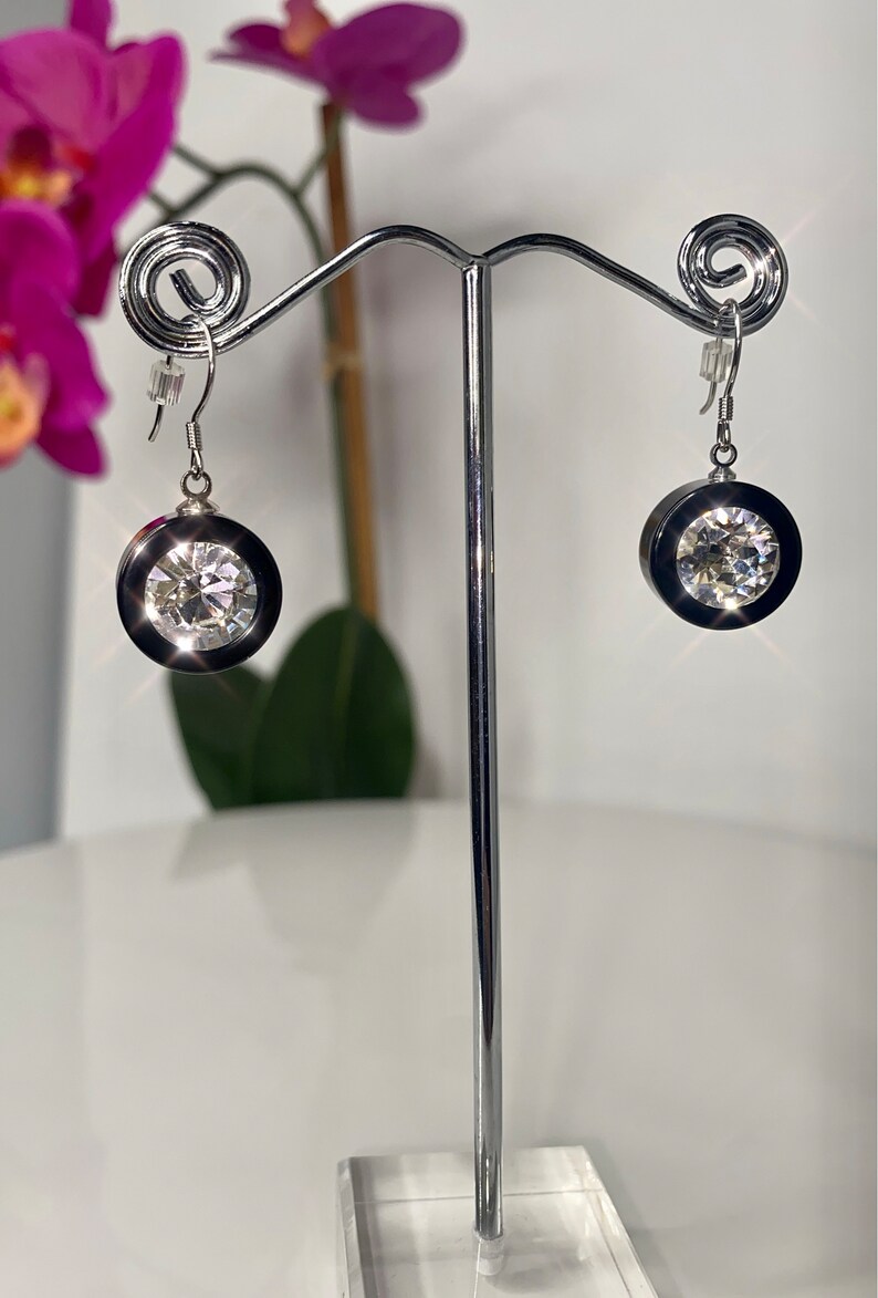 May include: A pair of silver earrings with a large, clear crystal stone set in a black bezel. The earrings are hanging from a silver stand.