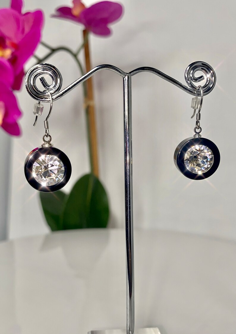 May include: A pair of silver dangle earrings with a black and clear crystal design. The earrings are hanging from a silver stand.