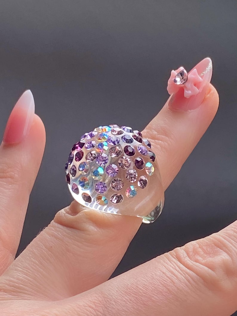 May include: A clear ring with a dome-shaped top covered in iridescent purple and pink rhinestones.