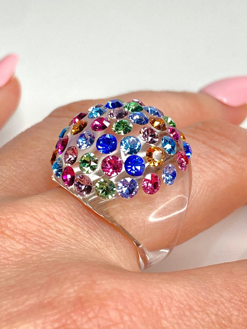 May include: A clear plastic ring with a dome-shaped top covered in colorful rhinestones. The rhinestones are in various shades of blue, green, pink, red, and orange.