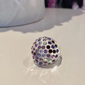 May include: A clear ring with a round, multi-colored design. The ring is covered in small, iridescent purple, blue, and white rhinestones.