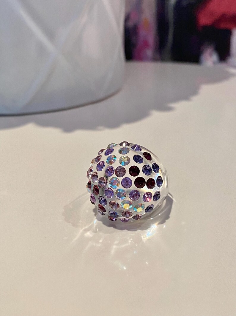 May include: A clear ring with a round, multi-colored design. The ring is covered in small, iridescent purple, blue, and white rhinestones.