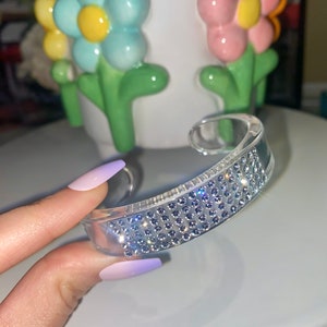 May include: Clear acrylic cuff bracelet with a row of small, sparkling rhinestones. The bracelet is held in front of a white ceramic vase decorated with colorful, three-dimensional flowers.