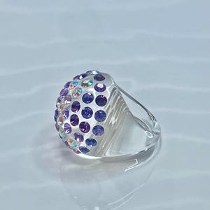 May include: A clear ring with a dome-shaped top covered in iridescent purple and pink rhinestones.