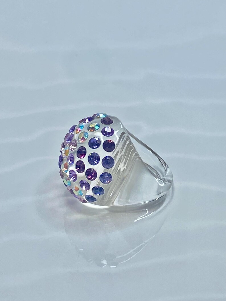 May include: A clear ring with a dome-shaped top covered in iridescent purple and pink rhinestones.