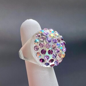 May include: A clear ring with a large, round, multi-colored crystal top. The crystal is set with a variety of iridescent purple, pink, and blue stones.