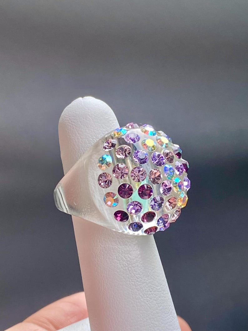 May include: A clear ring with a large, round, multi-colored crystal top. The crystal is set with a variety of iridescent purple, pink, and blue stones.