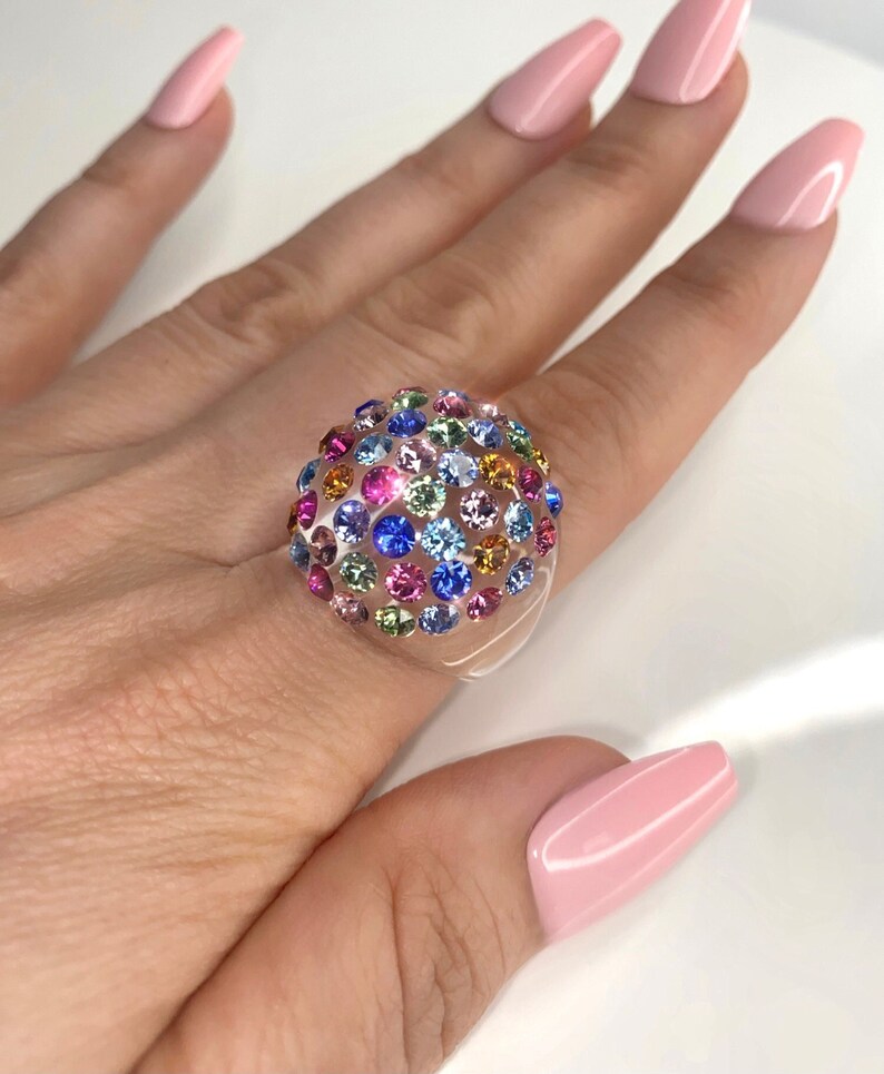May include: A clear ring with a dome-shaped top covered in colorful rhinestones. The rhinestones are in various shades of blue, green, pink, orange, and yellow.