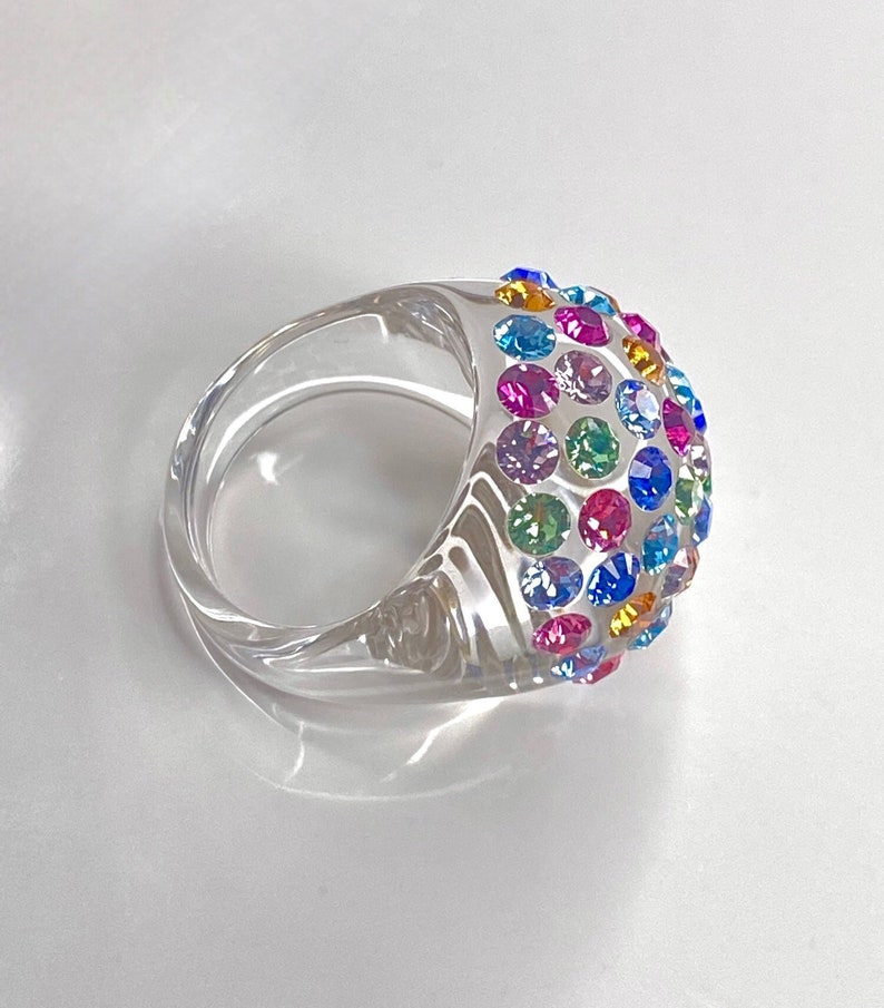 May include: A clear plastic ring with a dome-shaped top covered in colorful rhinestones. The rhinestones are in a variety of colors, including pink, blue, green, and purple.