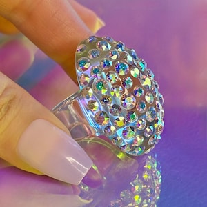 May include: A clear, oval ring covered in iridescent rhinestones. The ring's transparent band supports the array of colourful stones. The ring is held up against a reflective, iridescent surface, creating a shimmering effect.