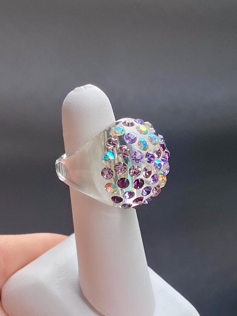 May include: A clear ring with a dome-shaped top covered in iridescent purple and blue rhinestones.
