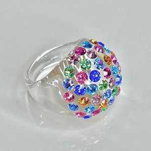 May include: A clear plastic ring with a dome-shaped top covered in colorful rhinestones. The rhinestones are in various shades of blue, green, pink, orange, and yellow.