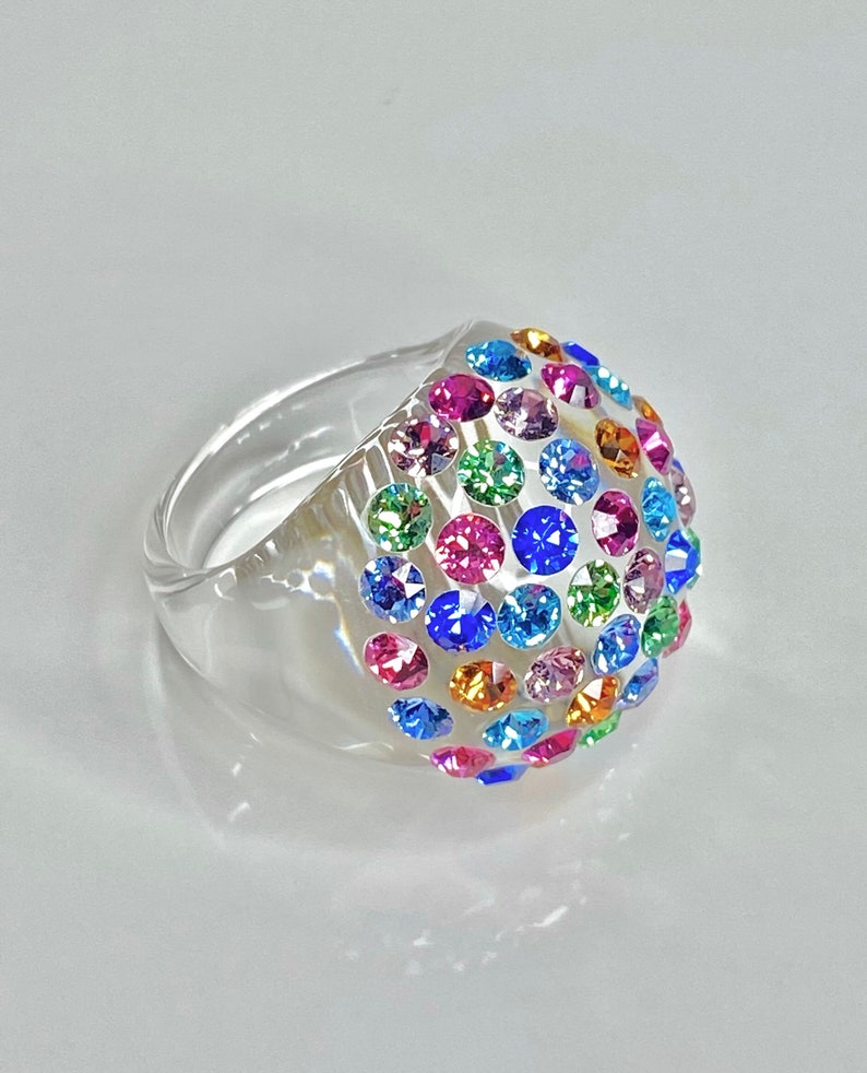 May include: A clear plastic ring with a dome-shaped top covered in colorful rhinestones. The rhinestones are in various shades of blue, green, pink, orange, and yellow.