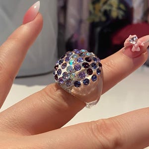 May include: A clear plastic ring with a dome-shaped top covered in iridescent purple, blue, and pink rhinestones.