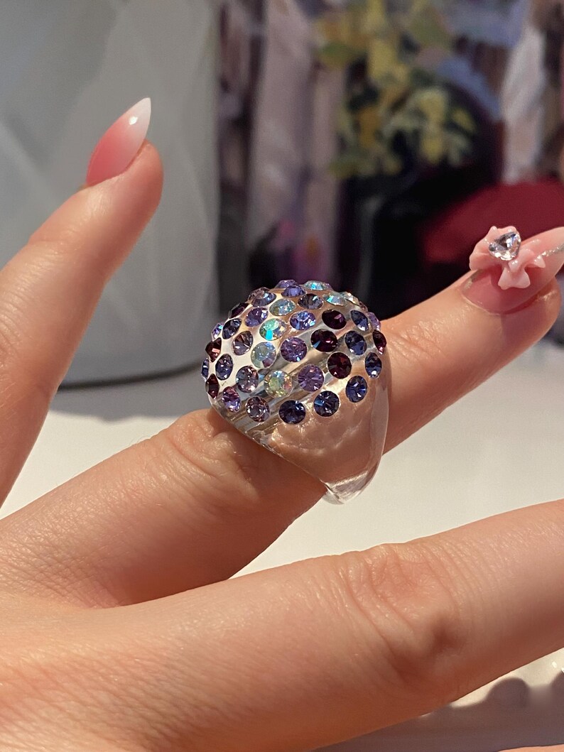 May include: A clear plastic ring with a dome-shaped top covered in iridescent purple, blue, and pink rhinestones.