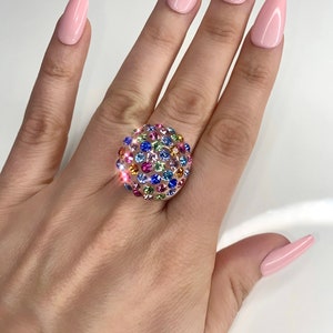 May include: A gold ring with a round, multi-colored stone setting. The stone is covered in small, colorful crystals.