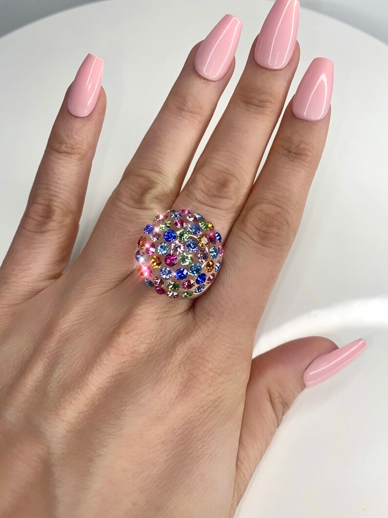 May include: A gold ring with a round, multi-colored stone setting. The stone is covered in small, colorful crystals.