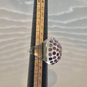May include: A clear glass ring with a dome of iridescent purple and pink rhinestones. The ring is sized on a ring sizer that shows the size is 5.5.