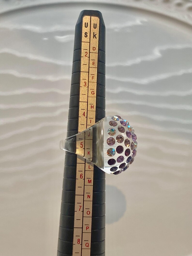 May include: A clear glass ring with a dome of iridescent purple and pink rhinestones. The ring is sized on a ring sizer that shows the size is 5.5.