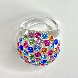May include: A clear plastic ring with a dome-shaped top covered in colorful rhinestones. The rhinestones are in various shades of blue, green, pink, orange, and purple.