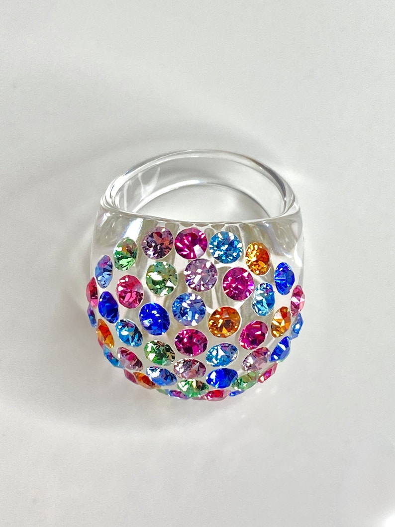 May include: A clear plastic ring with a dome-shaped top covered in colorful rhinestones. The rhinestones are in various shades of blue, green, pink, orange, and purple.