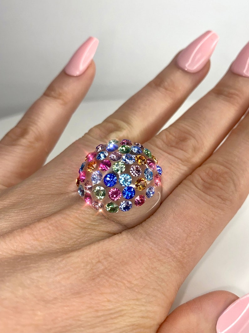 May include: A clear ring with a dome-shaped top covered in colorful rhinestones. The rhinestones are in shades of pink, blue, green, yellow, and orange.