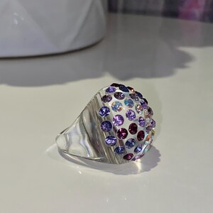 May include: A silver ring with a dome-shaped top covered in small, colorful gemstones. The gemstones are arranged in a gradient pattern, starting with light blue and transitioning to purple.