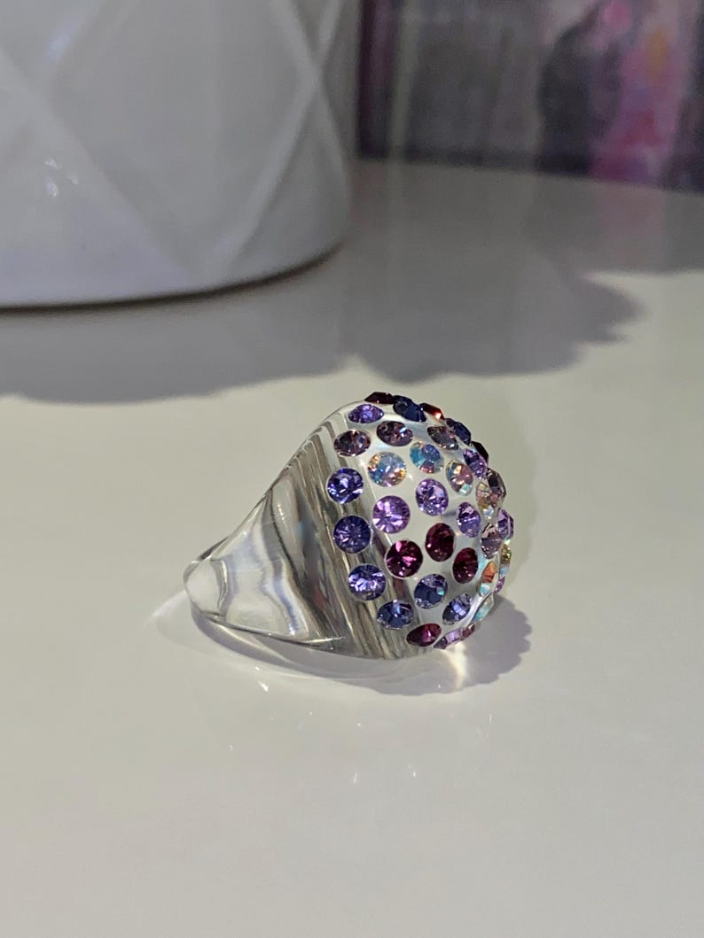 May include: A silver ring with a dome-shaped top covered in small, colorful gemstones. The gemstones are arranged in a gradient pattern, starting with light blue and transitioning to purple.