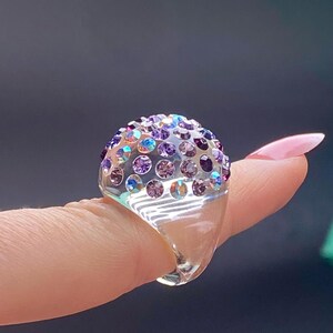 May include: A clear ring with a dome-shaped top covered in iridescent purple and blue rhinestones.