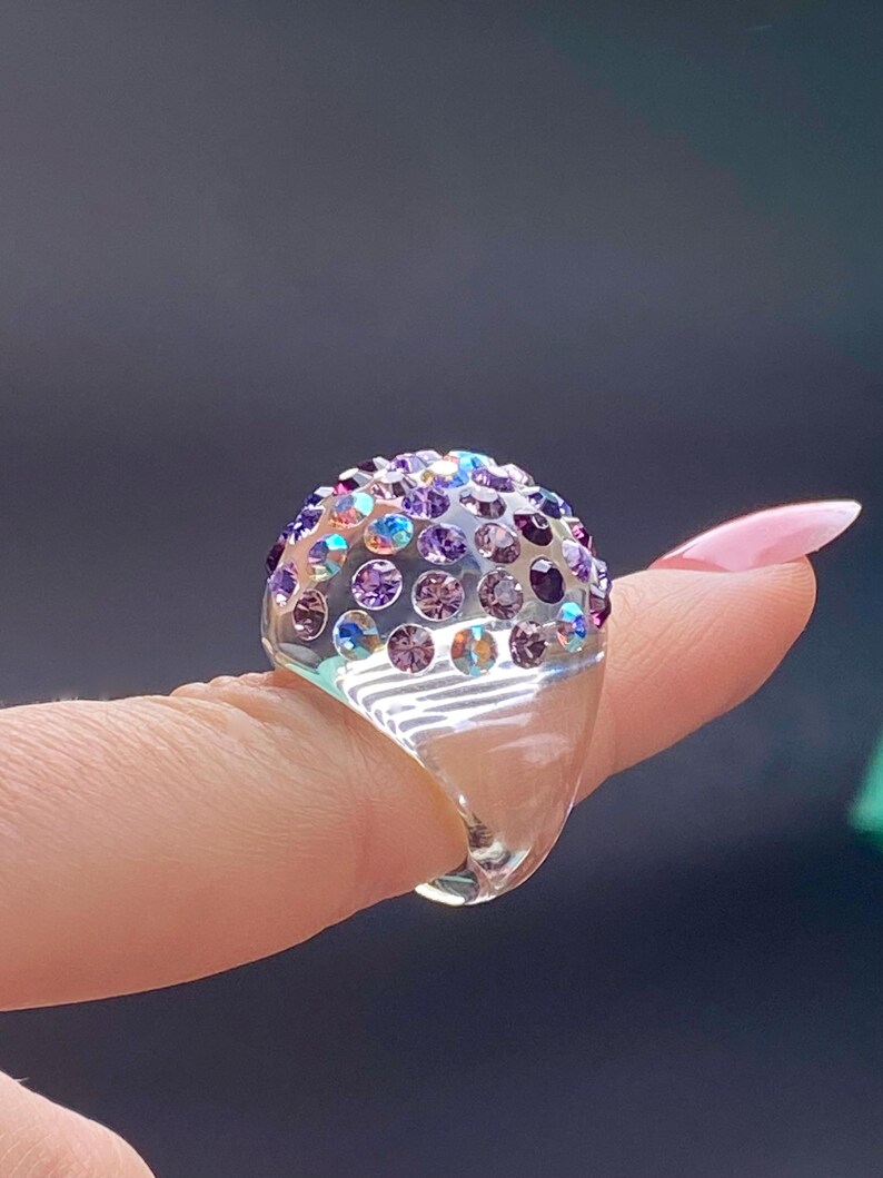 May include: A clear ring with a dome-shaped top covered in iridescent purple and blue rhinestones.