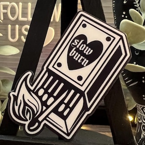 May include: Black and white sticker of a matchbox with a flame and the text "slow burn" on a small black easel.