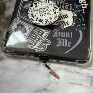 May include: A black phone case with a clear bumper, decorated with stickers and a pop socket. The stickers feature gothic-style text and illustrations. A small knife charm with the words "Killing it" hangs from the phone.