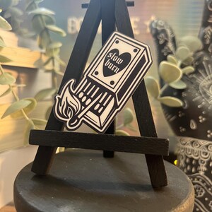 May include: A black and white sticker on a small black easel. The sticker is a matchbox with the text "slow burn" and a heart. The easel is on a black wooden surface.