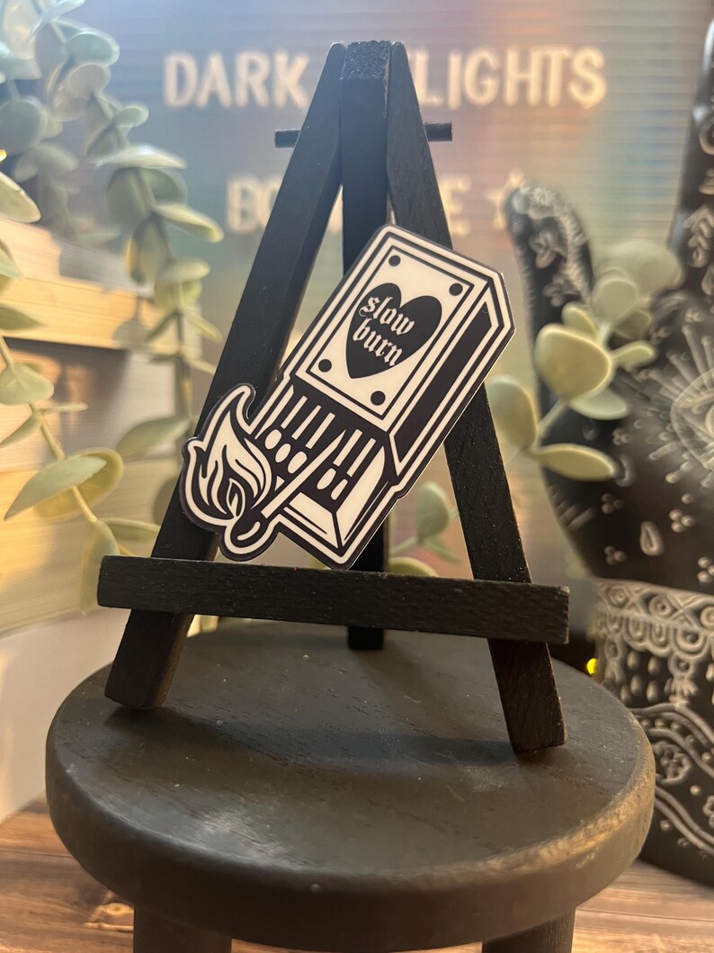 May include: A black and white sticker on a small black easel. The sticker is a matchbox with the text "slow burn" and a heart. The easel is on a black wooden surface.