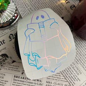May include: Holographic sticker featuring a ghost-like figure with a rectangular head and flowing robes. The sticker has a pearlescent sheen, reflecting colors like blue, pink, and yellow. The design is set against a backdrop of newspaper print.