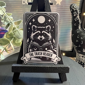 May include: A black and white sticker featuring a raccoon reading a book with the text "THE TRASH READER" on a white banner. The raccoon is sitting under a full moon with stars in the background.