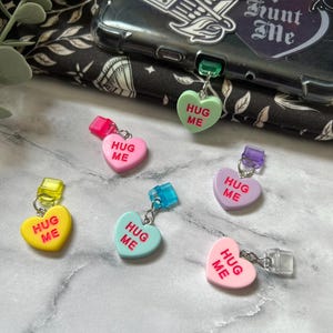 May include: Six heart-shaped charms with the text "HUG ME" in red lettering. The charms are in various colors: pink, yellow, blue, green, and purple. Each charm has a clear plastic ring and a metal loop for attaching to a bag or keychain.