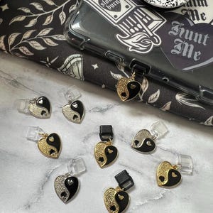 May include: Eight black and gold yin yang heart charms with a small heart in the center of each yin yang symbol. The charms are on a white marble surface.