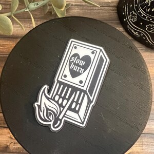 May include: Black and white sticker of a matchbox with a heart and the text "slow burn". The matchbox is on fire.